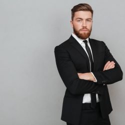 Confident young businessman in suit standing with arms folded isolated over gray background