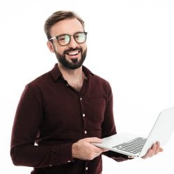 Portrait of a smiling handsome man in eyeglasses holding laptop computer and looking at camera isolated over white background